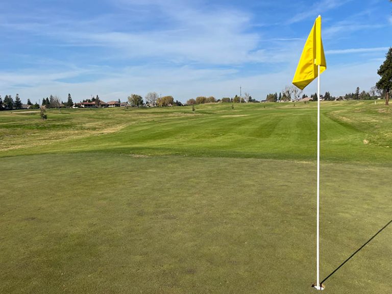 Yellow flagstick on a golf green looking back toward a wide rolling fairway and distant homes.